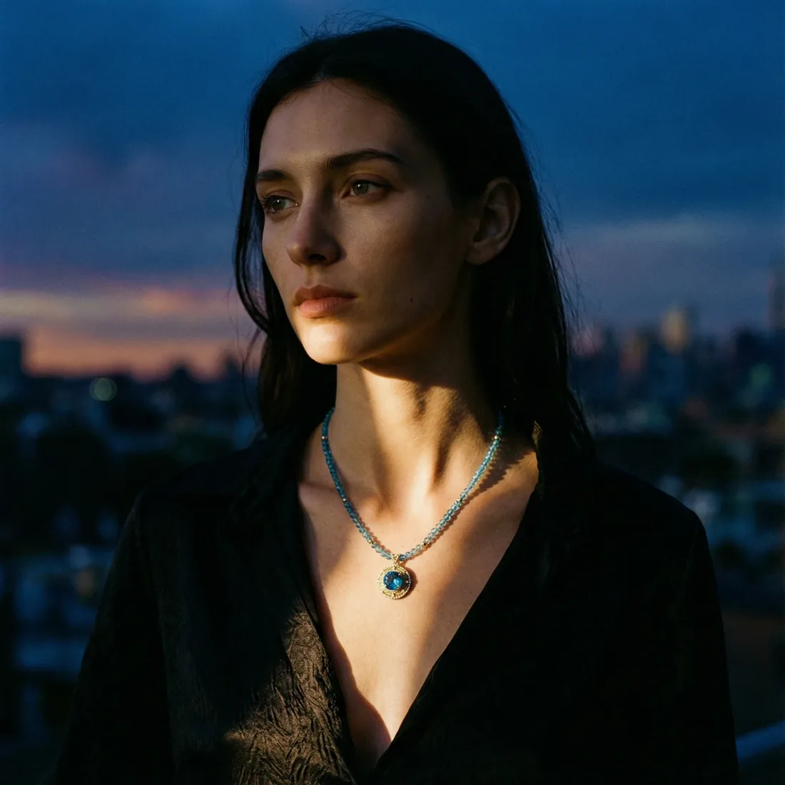 Woman wearing a gold and blue gemstone pendant necklace at dusk with the New York City skyline behind her