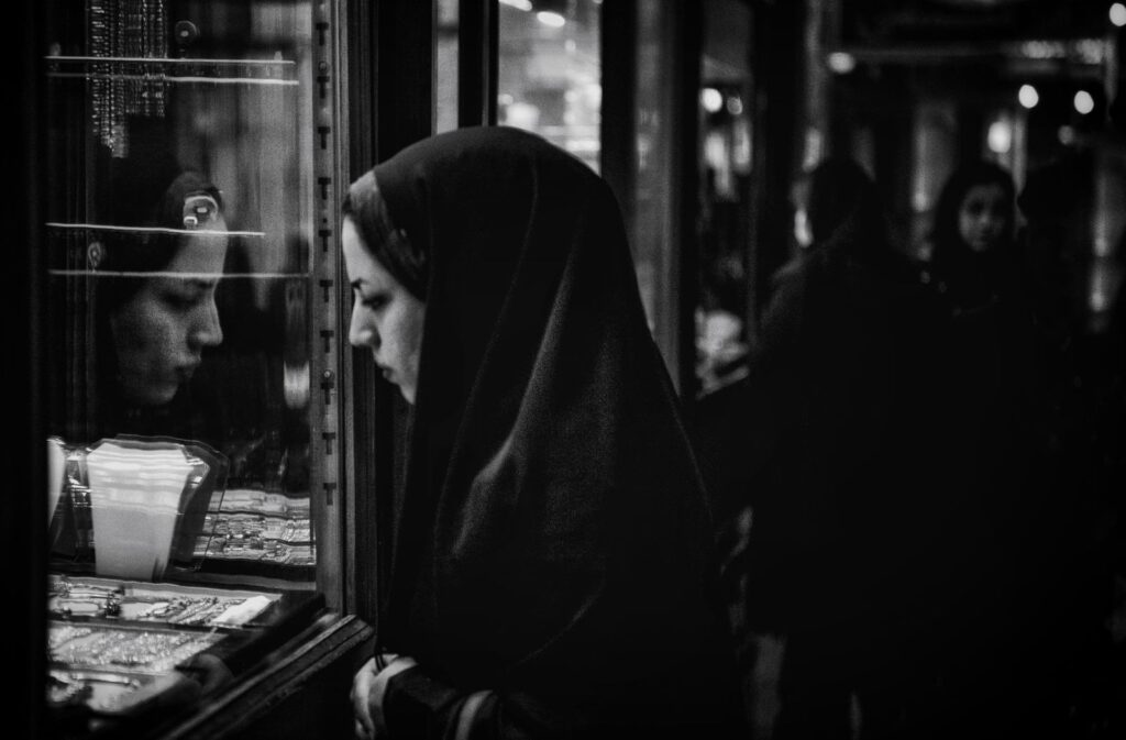 A woman studying a lit jewelry display case at night, her own reflection visible in the glass