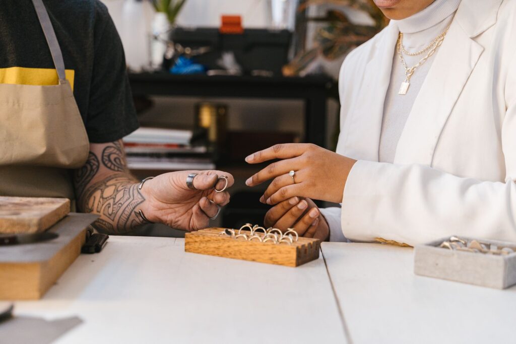 A jeweler in a workshop apron showing a tray of rings to a customer across the bench, the moment of handing something over
