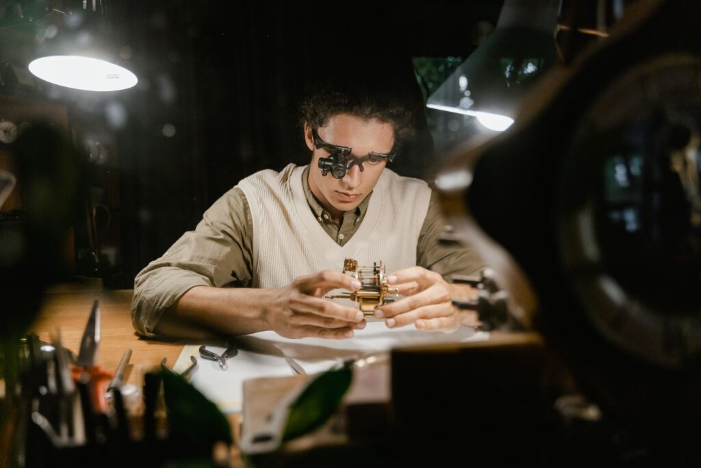 A watchmaker at a lit bench examining a movement through a headband magnifier, diagnosing one component at a time